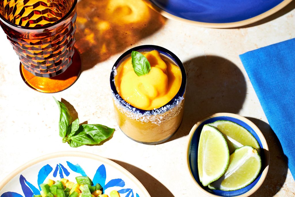 frozen mango margarita in a glass garnished with a basil leaf with a salt rim with lime wedges in front with tortilla chips in the back on a neutral background