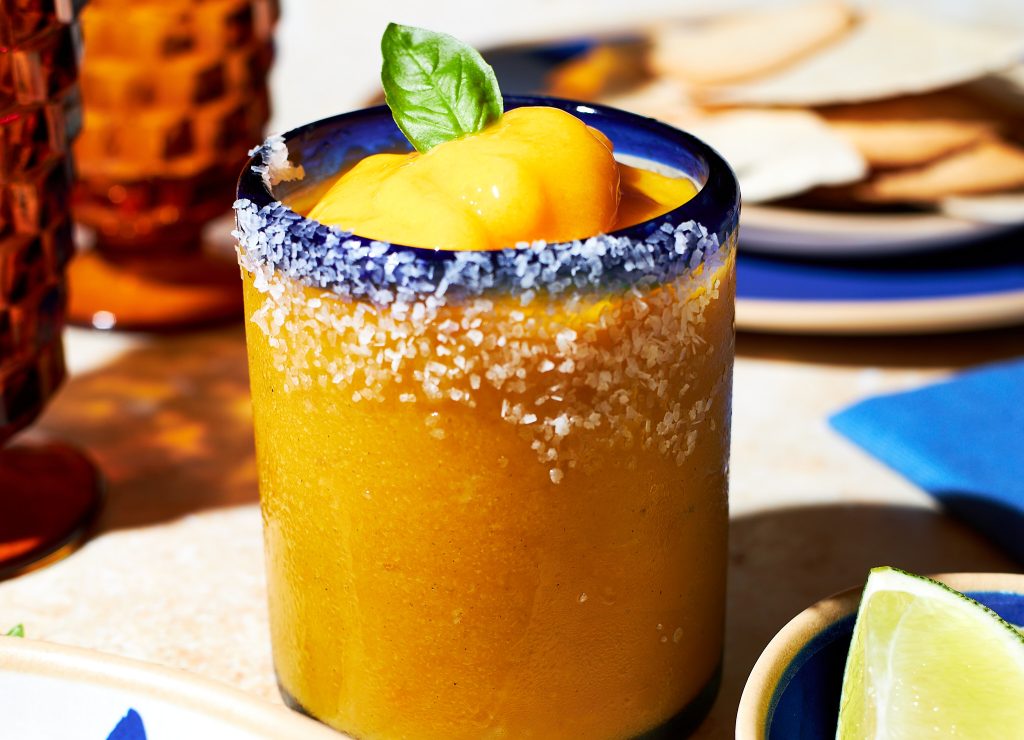 frozen mango margarita in a glass garnished with a basil leaf with a salt rim with lime wedges in front with tortilla chips in the back on a neutral background