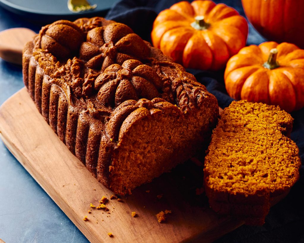 pumpkin loaf made with nordicware pumpkin wheat loaf pan on a blue background with cinnamon, pumpkins, forks, plates, and napkins