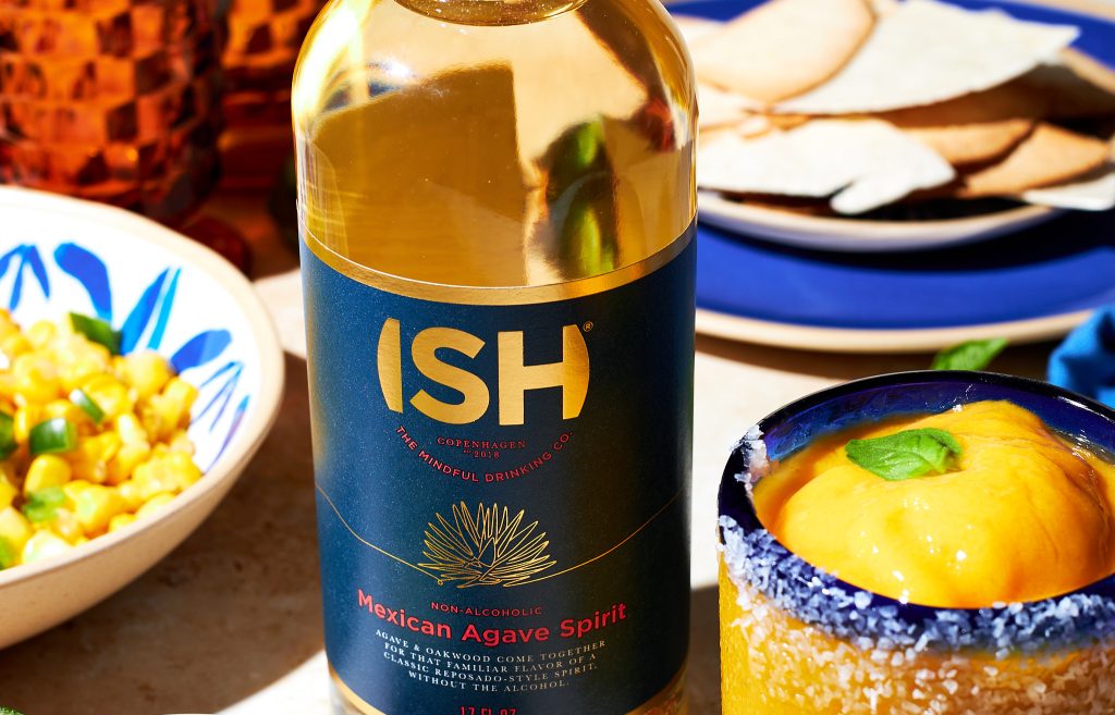 ish non-alcoholic mexican agave spirit. frozen mango margarita in a glass garnished with a basil leaf with a salt rim with lime wedges in front with tortilla chips in the back on a neutral background