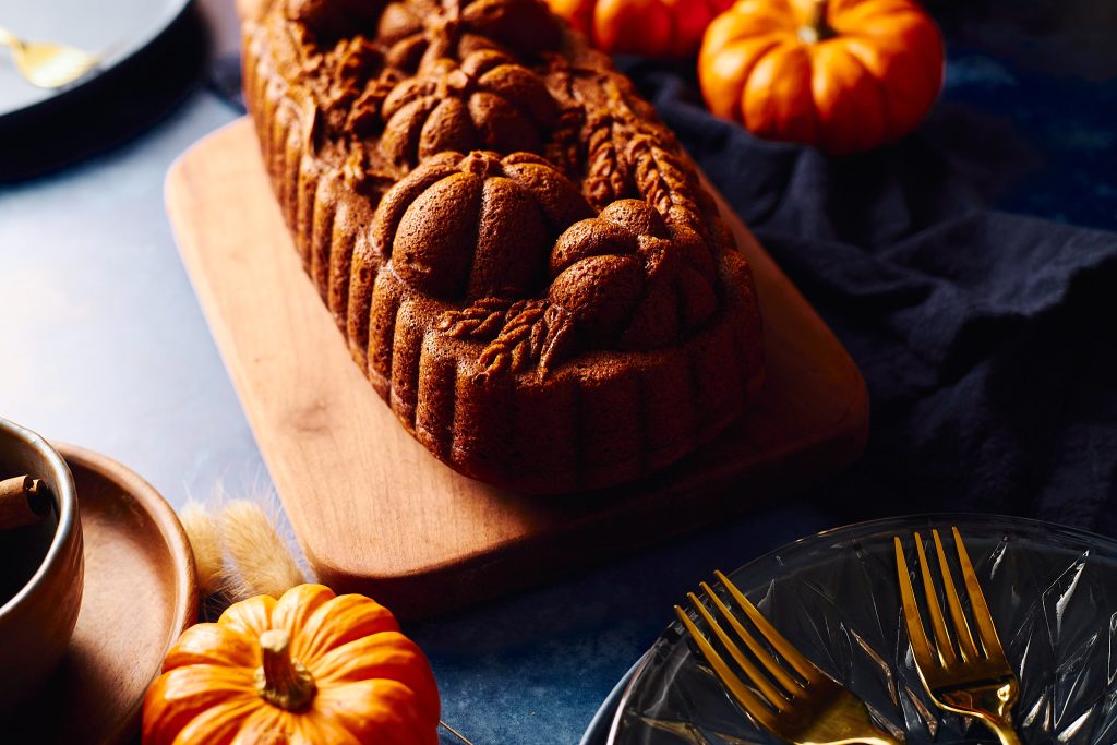 pumpkin loaf made with nordicware pumpkin wheat loaf pan on a blue background with cinnamon, pumpkins, forks, plates, and napkins