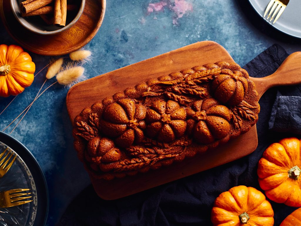 pumpkin loaf made with nordicware pumpkin wheat loaf pan on a blue background with cinnamon, pumpkins, forks, plates, and napkins