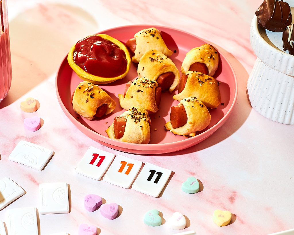 vegetarian pigs in a blanket on a pink plate with ketchup for dipping with valentine's candy and rummikub int he background on a light pink surface