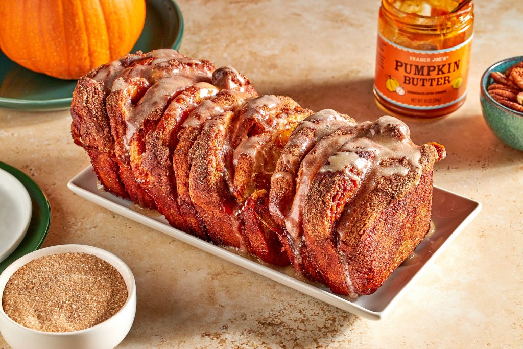 Cinnamon pull apart bread made with biscuits and cinnamon rolls and trader joe's pumpkin butter with a bowl of cinnamon sugar on a neutral background with plates and a pumpkin in the background