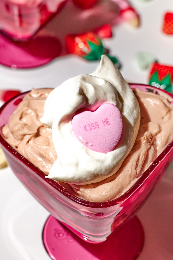 chocolate cheesecake mousse in a heart shaped dessert coupe with whipped cream on top on a white background with conversation hearts candy