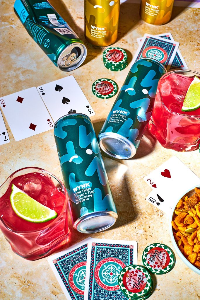 Wynk mocktail in a glass with wynk lime twist selszer beside it with playing cards, poker chips, snack mix in a bowl, on a neutral background