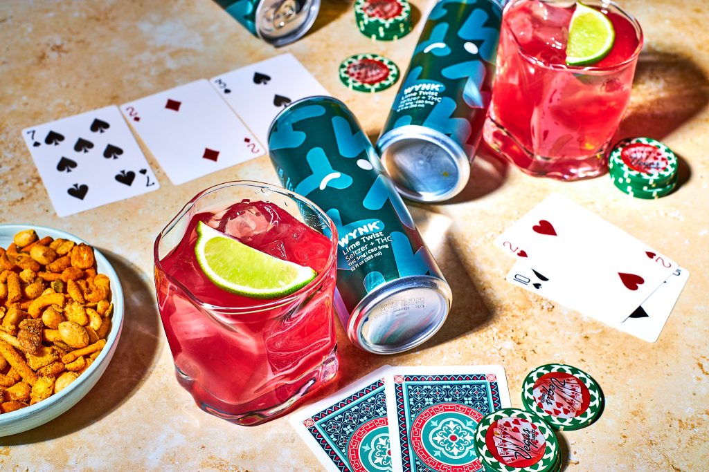 Wynk mocktail in a glass with wynk lime twist selszer beside it with playing cards, poker chips, snack mix in a bowl, on a neutral background