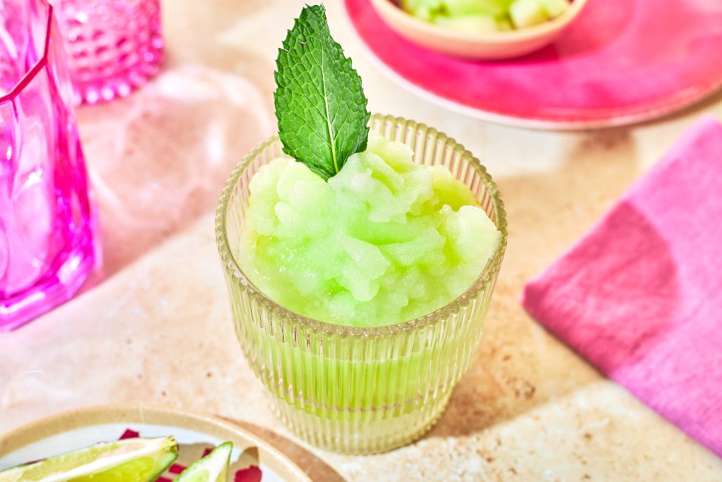 honeydew frozen margarita mocktail in a ribbed glass with a mint leaf garnish with mint, limes, and honeydew in bowls in the background with a pink napkin and a pink glass on a neutral background