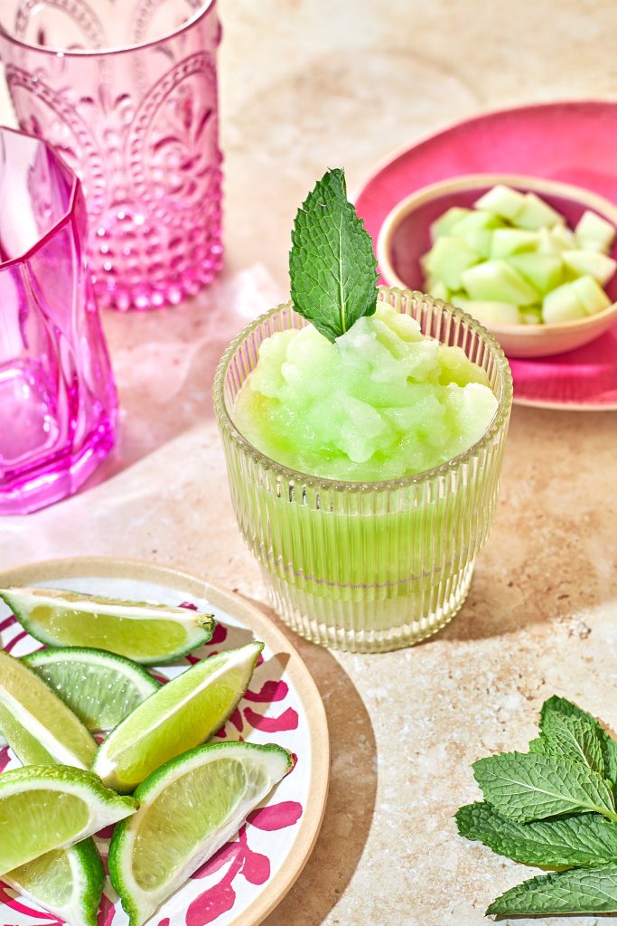 honeydew frozen margarita mocktail in a ribbed glass with a mint leaf garnish with mint, limes, and honeydew in bowls in the background with a pink napkin and a pink glass on a neutral background