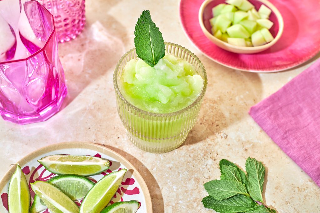 honeydew frozen margarita mocktail in a ribbed glass with a mint leaf garnish with mint, limes, and honeydew in bowls in the background with a pink napkin and a pink glass on a neutral background