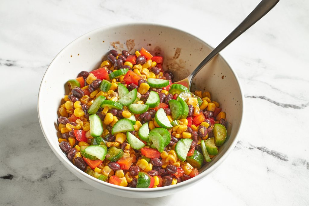 cucumbers, red bell peppers, black beans, corn, in a taco sauce, taco dense bean salad recipe in a bowl on a neutral surface